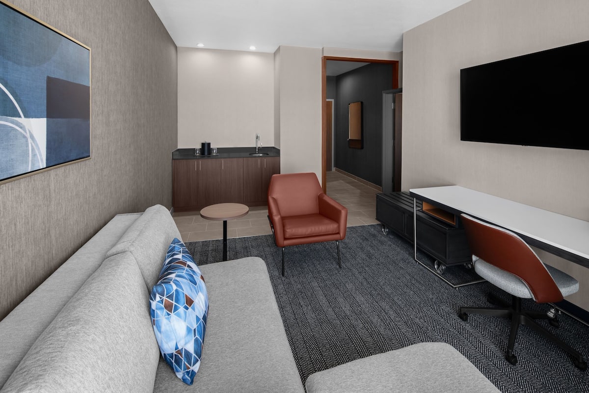 Ett modernt hotellrum med en grå soffa, en brun läderfåtölj, ett vitt skrivbord och en platt-TV som är monterad på väggen.