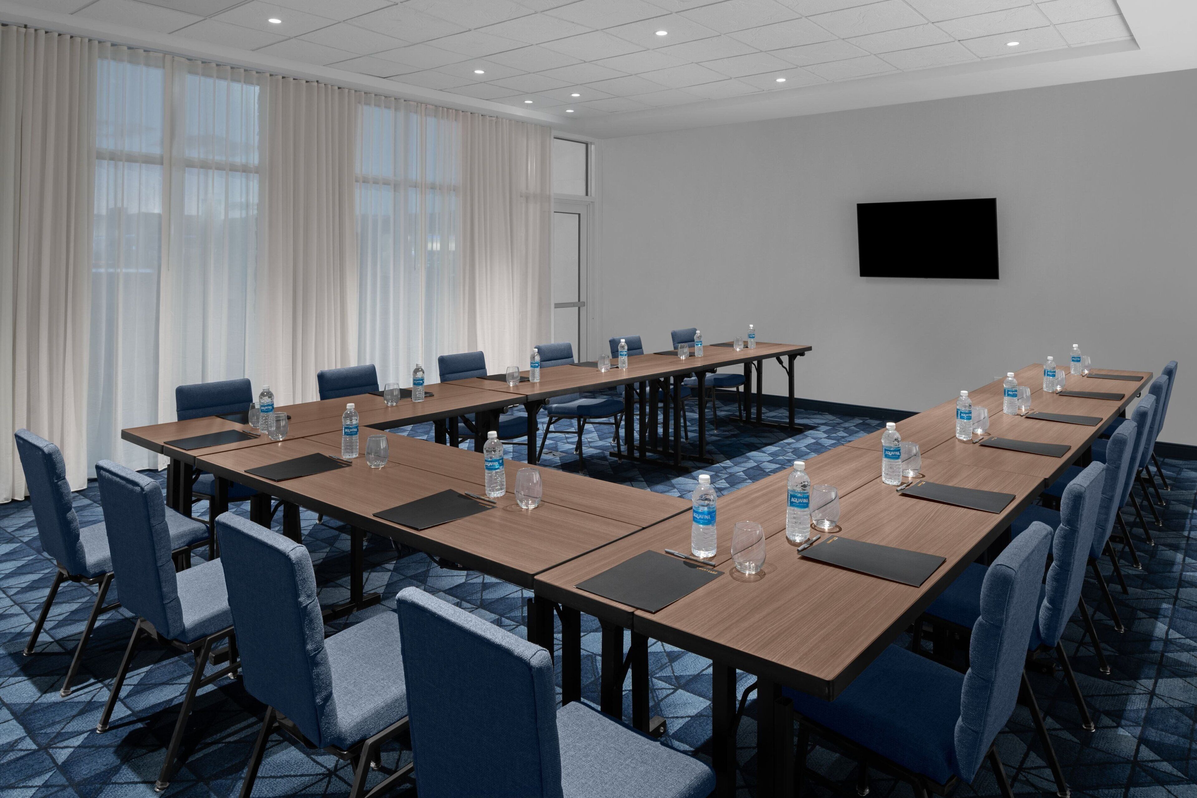 A conference room with long tables, blue chairs, water bottles, and a TV on the wall.