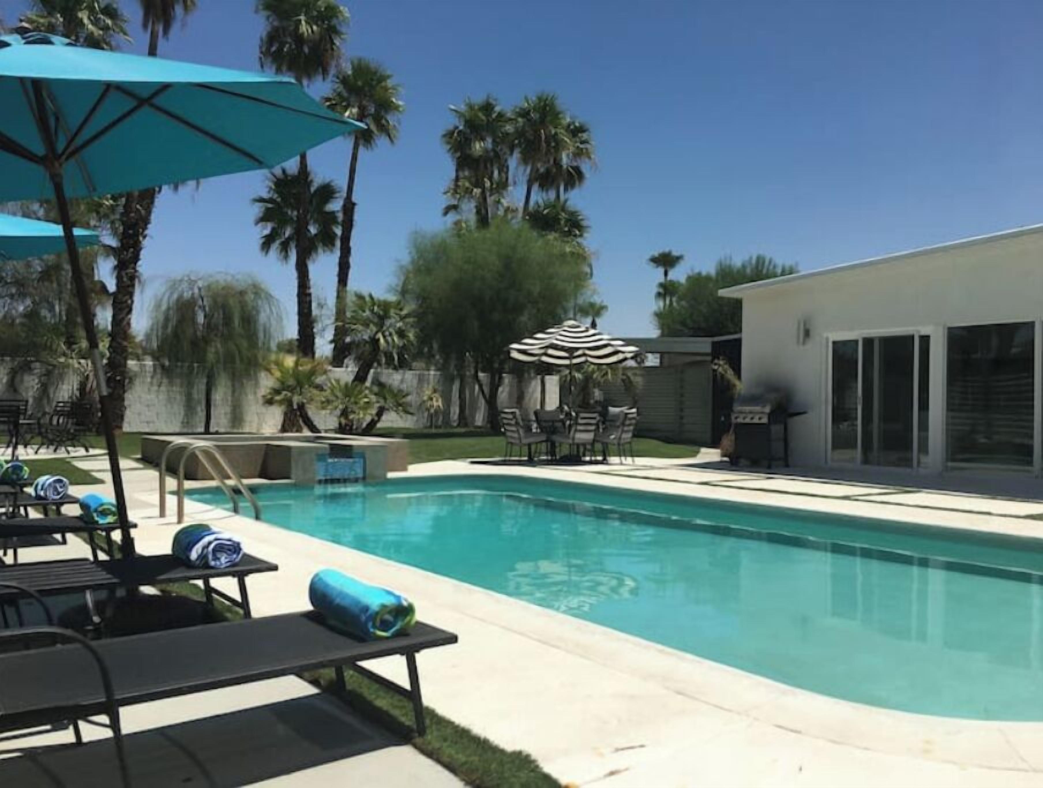 A swimming pool with lounge chairs and umbrellas, surrounded by palm trees and a modern house.