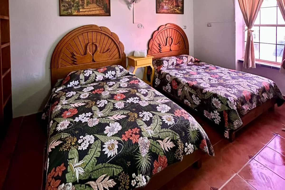 Une pièce comprenant deux lits en bois recouverts de couvre-lits à motifs floraux, un ventilateur sur pied et deux tableaux accrochés au mur.