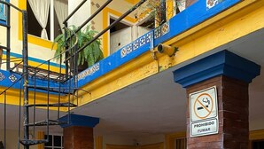Entrada de un edificio con un letrero de prohibido fumar, paredes pintadas de amarillo y azul, y mobiliario exterior con sillas.