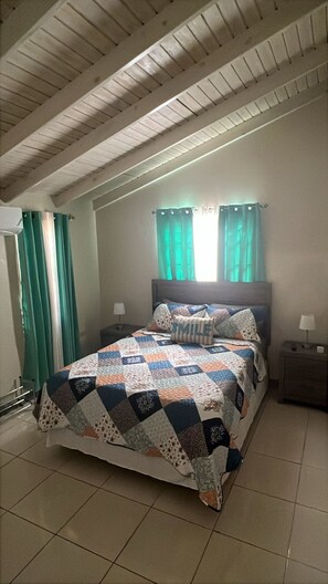 Un dormitorio con techo inclinado, una cama con colcha, dos lámparas de noche y una ventana con cortinas.