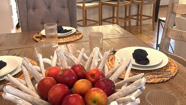 Een eettafel met een fruitmand, houten stoelen en een moderne keuken op de achtergrond.