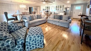 Een ruime woonkamer met een hoekbank, een salontafel en een gestreept poef. Op de achtergrond is een eethoek met een tafel en stoelen te zien.