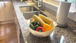 Een keuken met granieten werkbladen, een roestvrijstalen spoelbak, een schaal groenten en een raam met uitzicht op groen.