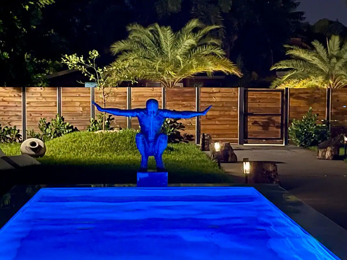 Ein Poolbereich bei Nacht mit einer blauen Statue in der Mitte, umgeben von Holzzaun und Palmen.