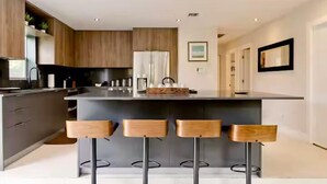A modern kitchen with a central island, bar stools, wooden cabinets, and a refrigerator. - Tree House (Fort Lauderdale)