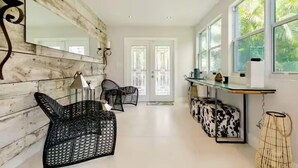A spacious room with a wooden accent wall, a large mirror, two black wicker chairs, a glass table, and a view of greenery through large windows. - Tree House (Fort Lauderdale)