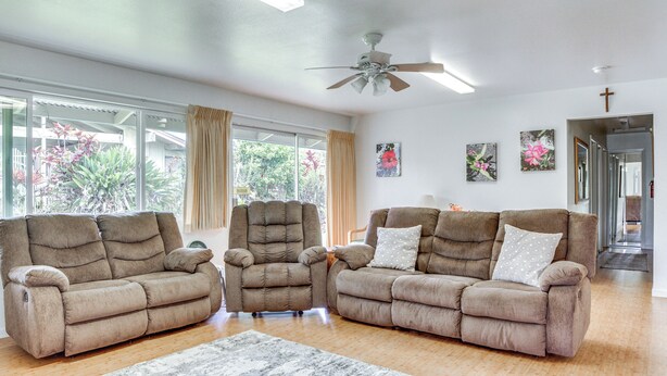 A spacious living room with two brown sofas, a ceiling fan, and large windows offering a view of greenery outside. - Sunny Hilo Home w/ Game Room: Walk to Golf Course! (Hilo)