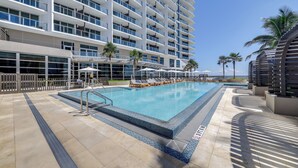 Una piscina con sillas de descanso y sombrillas, rodeada por un edificio alto con balcones y palmeras.
