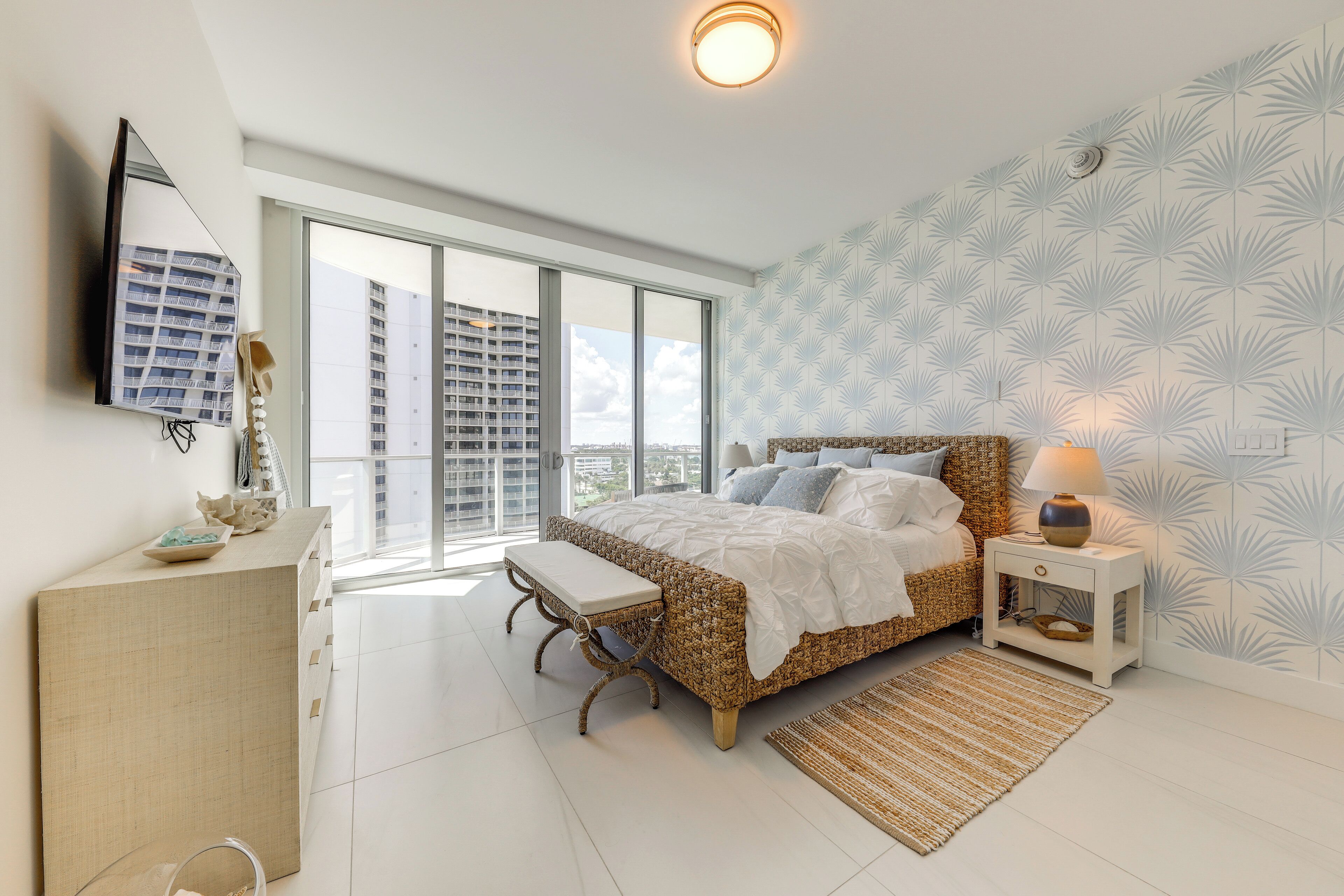 A bedroom with a large window, a bed, a bedside table, a lamp, and a wall-mounted TV.
