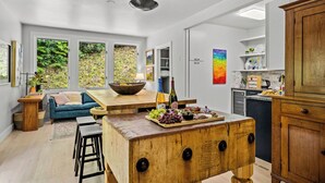 A modern kitchen with a wooden island, bar stools, and a dining area with a blue sofa. There are large windows with a view of greenery outside. - Luxe Mountain Home: Hot Tub, Sauna, Firepit, Games (Black Mountain)