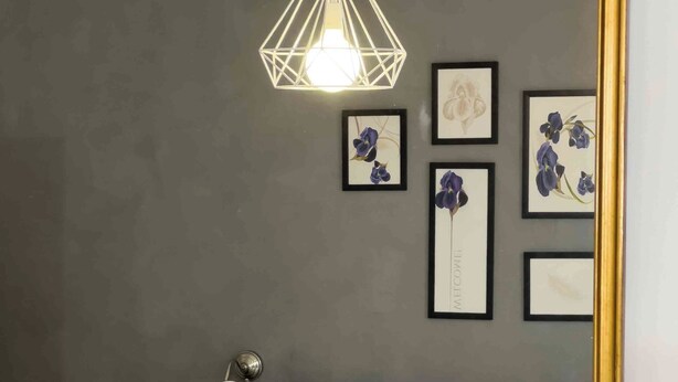 Een slaapkamer met een grote spiegel, een hangende geometrische lamp en een bed met witte lakens. Op de muur hangt een lijst met bloemistisch kunstwerk.