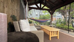 Un espacio de estar techado al aire libre con mobiliario de mimbre, una hamaca y vistas a un barrio residencial.