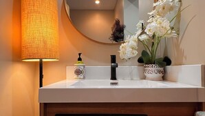 Un baño con un lavabo blanco, un espejo redondo, una lámpara de pared y un florero con flores blancas.