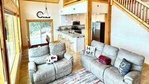 A spacious living area with a high ceiling, wooden beams, a balcony, and a kitchen in the background.