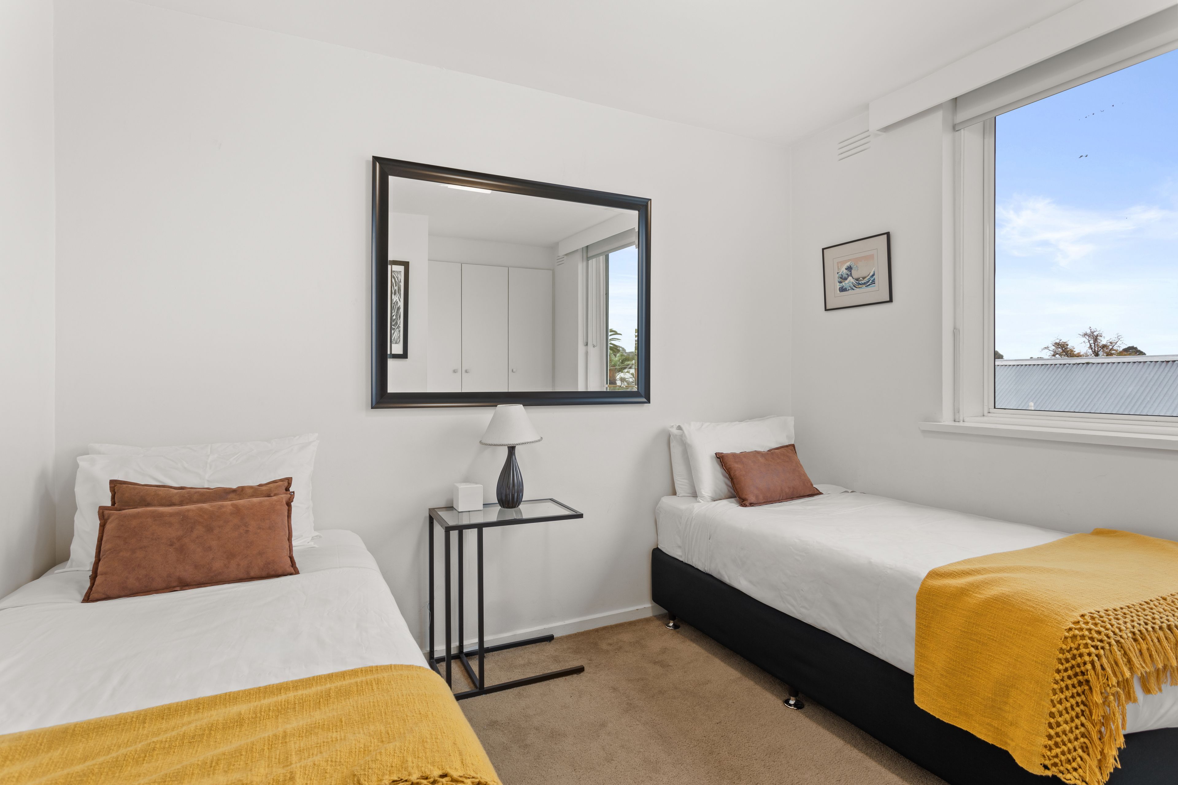 A bedroom with two beds, a bedside table with a lamp, a mirror, and a window with a view of the sky and trees.