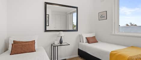 A bedroom with two beds, a bedside table with a lamp, a mirror, and a window with a view of the sky and trees.