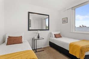 A bedroom with two beds, a bedside table with a lamp, a mirror, and a window with a view of the sky and trees.