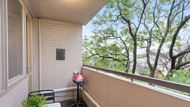 Un balcon avec une table, une chaise, un pot de fleurs, un barbecue et une vue sur les arbres et les bâtiments.