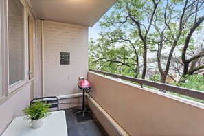 Un balcón con una mesa, una silla, una planta en maceta, una parrilla y vistas a árboles y edificios.