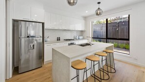 Una cocina moderna con armarios blancos, un refrigerador de acero inoxidable, una isla central con taburetes y un amplio ventanal con vistas al jardín trasero.