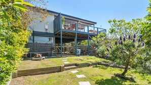 A modern two-story house with a covered outdoor area, a garden with a tree, and a clear sky.