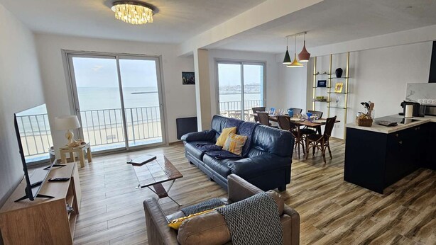 Amplia sala de estar con sofá, mesa de centro y mesa de comedor. Cuenta con una cocina con encimera y armarios. Amplias ventanas ofrecen vistas al exterior.