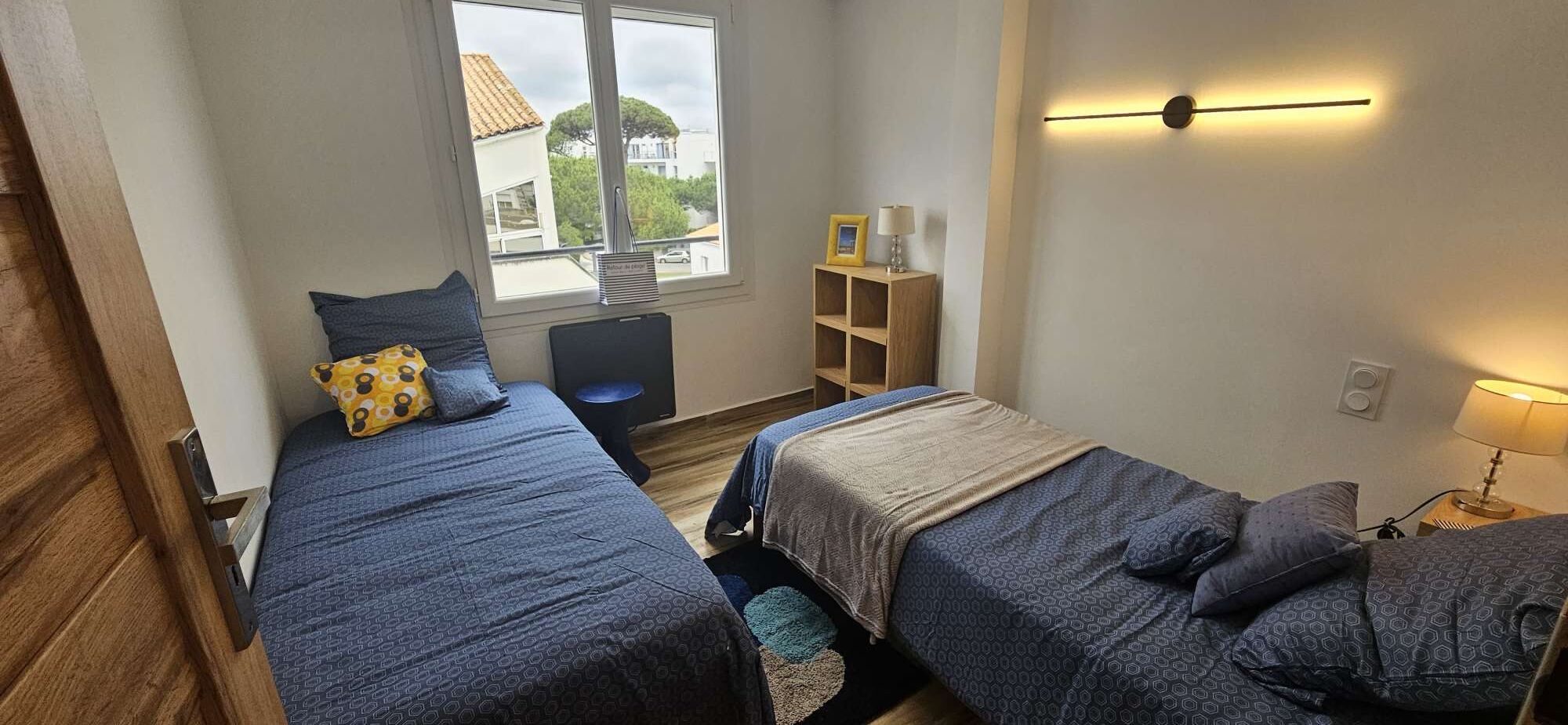 Una habitación con dos camas, un escritorio con silla, una ventana y una estantería con lámpara.