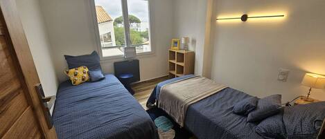 Una habitación con dos camas, un escritorio con silla, una ventana y una estantería con lámpara.