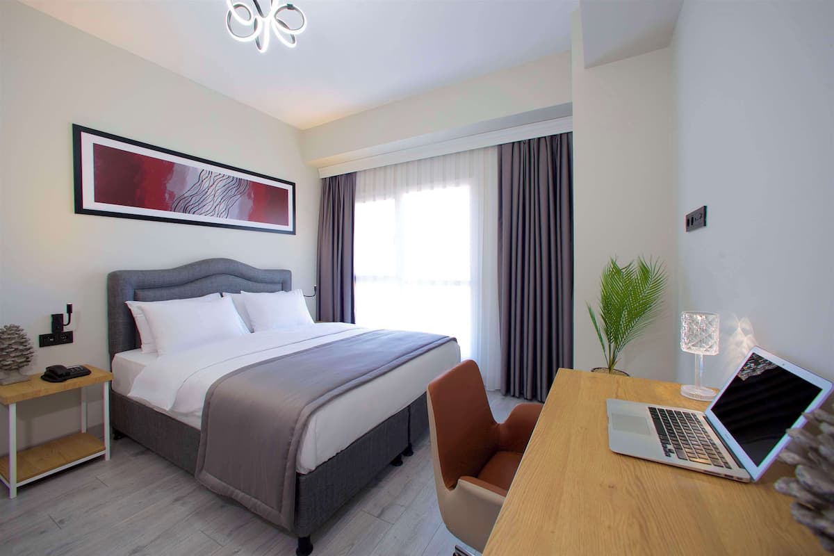 Ein Hotelzimmer mit einem Bett, einem Schreibtisch mit einem Laptop, einem Sessel, einem Nachttisch mit einer Lampe und einem großen Fenster mit Vorhängen.