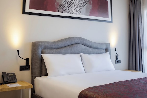 A neatly made bed with a grey upholstered headboard, white pillows, and a red patterned throw. There is a bedside table with a phone and a lamp, and a painting above the bed. - Best Western Plus Hotel & Residences Istanbul (Istanbul)