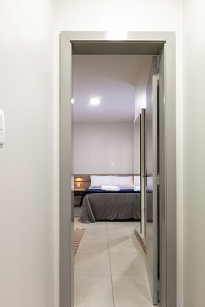 A narrow hallway leading to a bedroom with a bed, a nightstand, and a wall-mounted light. - MERANO GRAMADO - CENTRO 01 (Gramado)