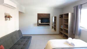 Moderni hotellihuone, jossa on sohva, valkoisella peitolla varustettu sänky, taulutelevisio, peili ja sisäänrakennetut hyllyt.