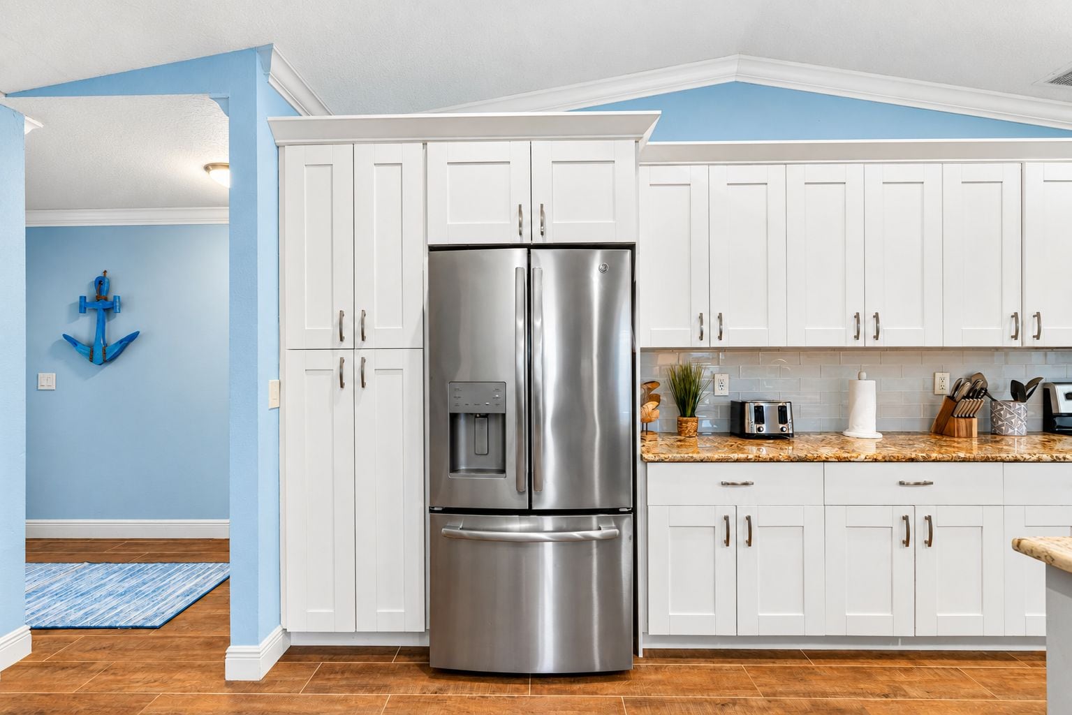 A kitchen with white cabinets, a stainless steel refrigerator, a granite countertop, and a blue wall with an anchor decoration.