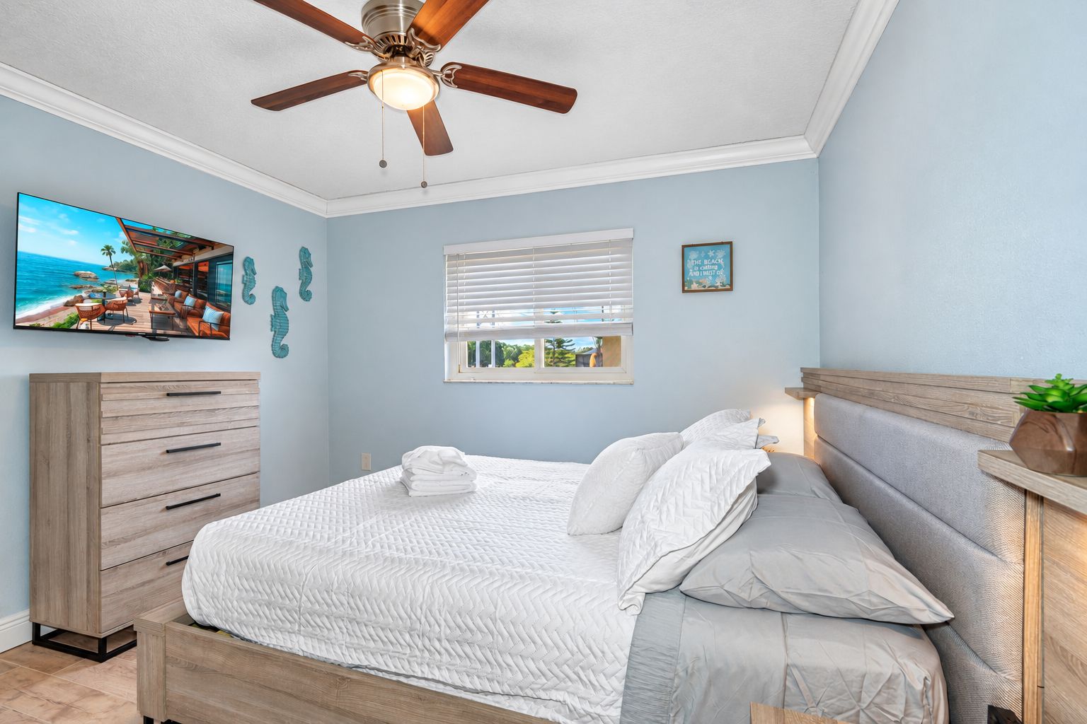 A bedroom with a bed, a ceiling fan, a dresser, a window with blinds, and a wall-mounted TV.