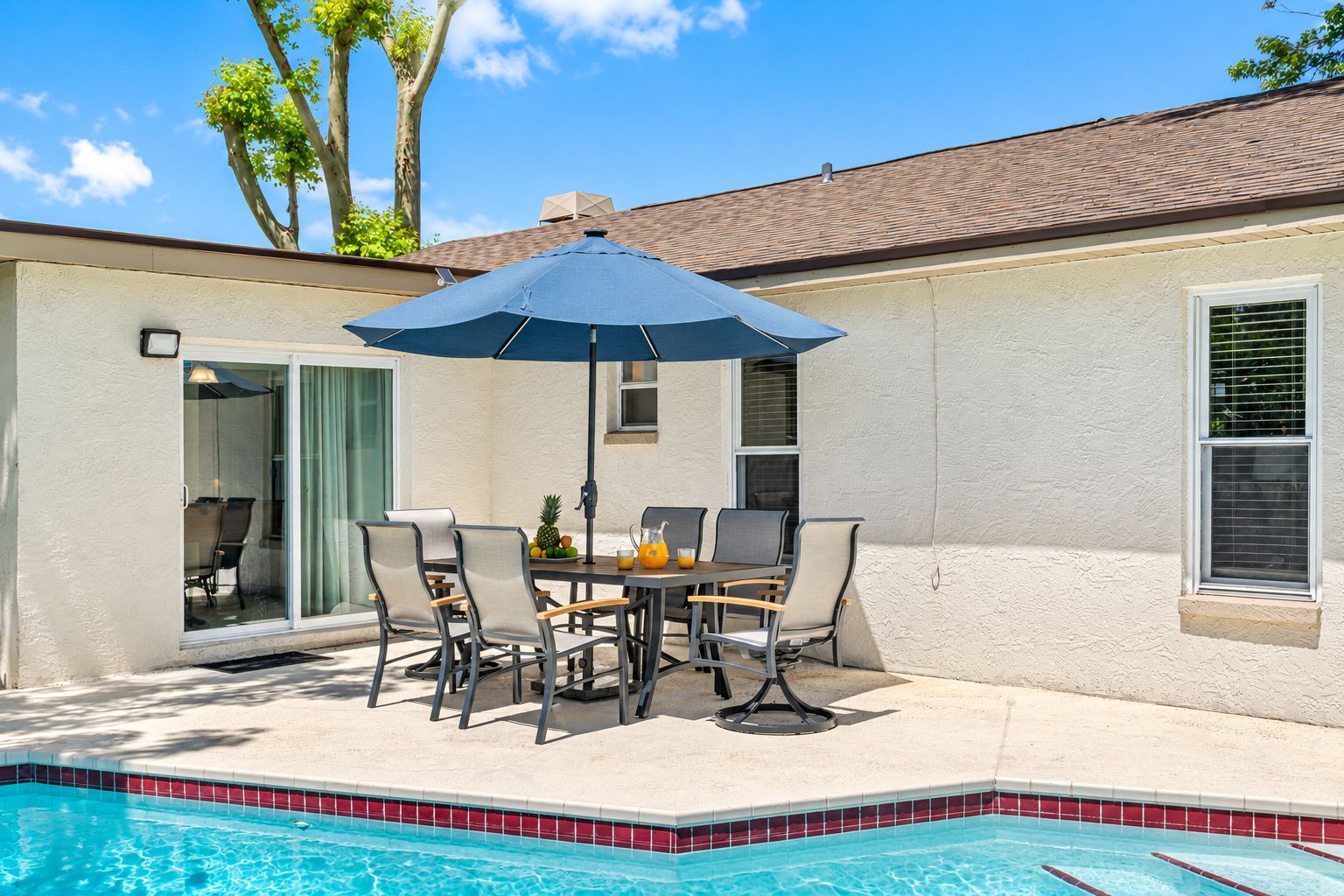 A poolside area with a table and chairs, a blue umbrella, and a view of the pool.