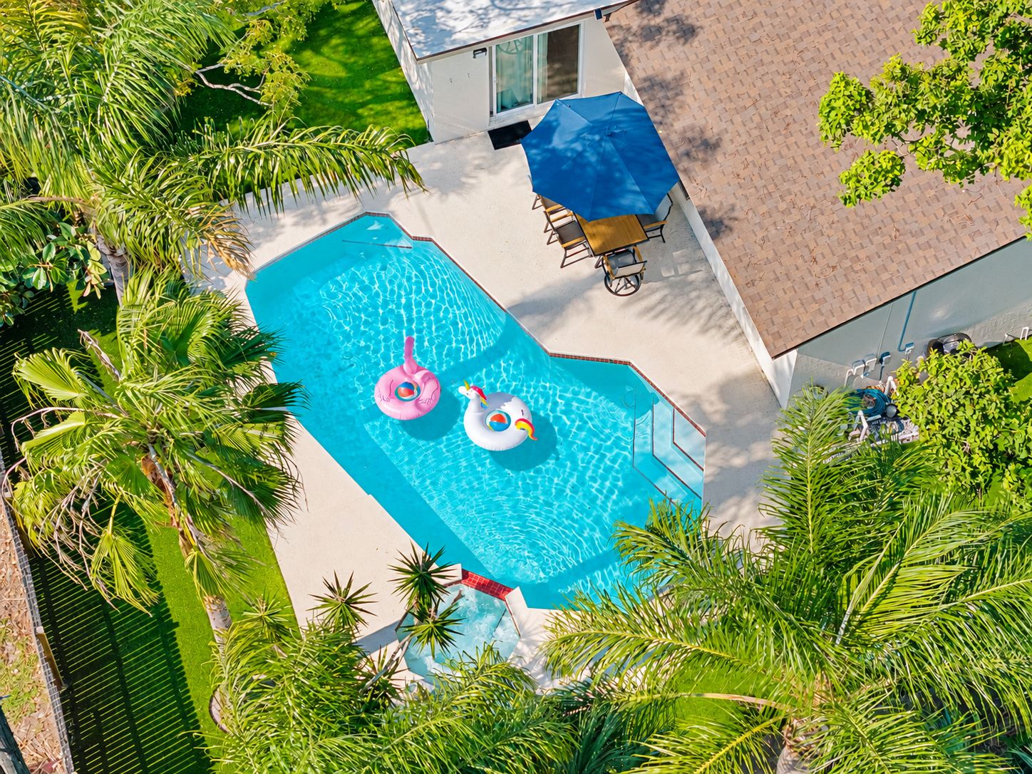 An aerial view of a swimming pool with two inflatable rings, surrounded by palm trees and a patio area with a table and chairs.