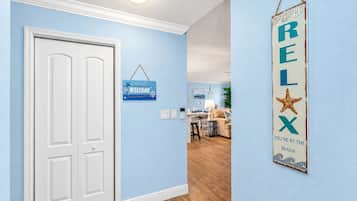 Un couloir avec une porte blanche, une pancarte indiquant « Bienvenue », une autre pancarte avec le mot « DÉTENTE », et un aperçu d’un salon comprenant une table à manger et des chaises.