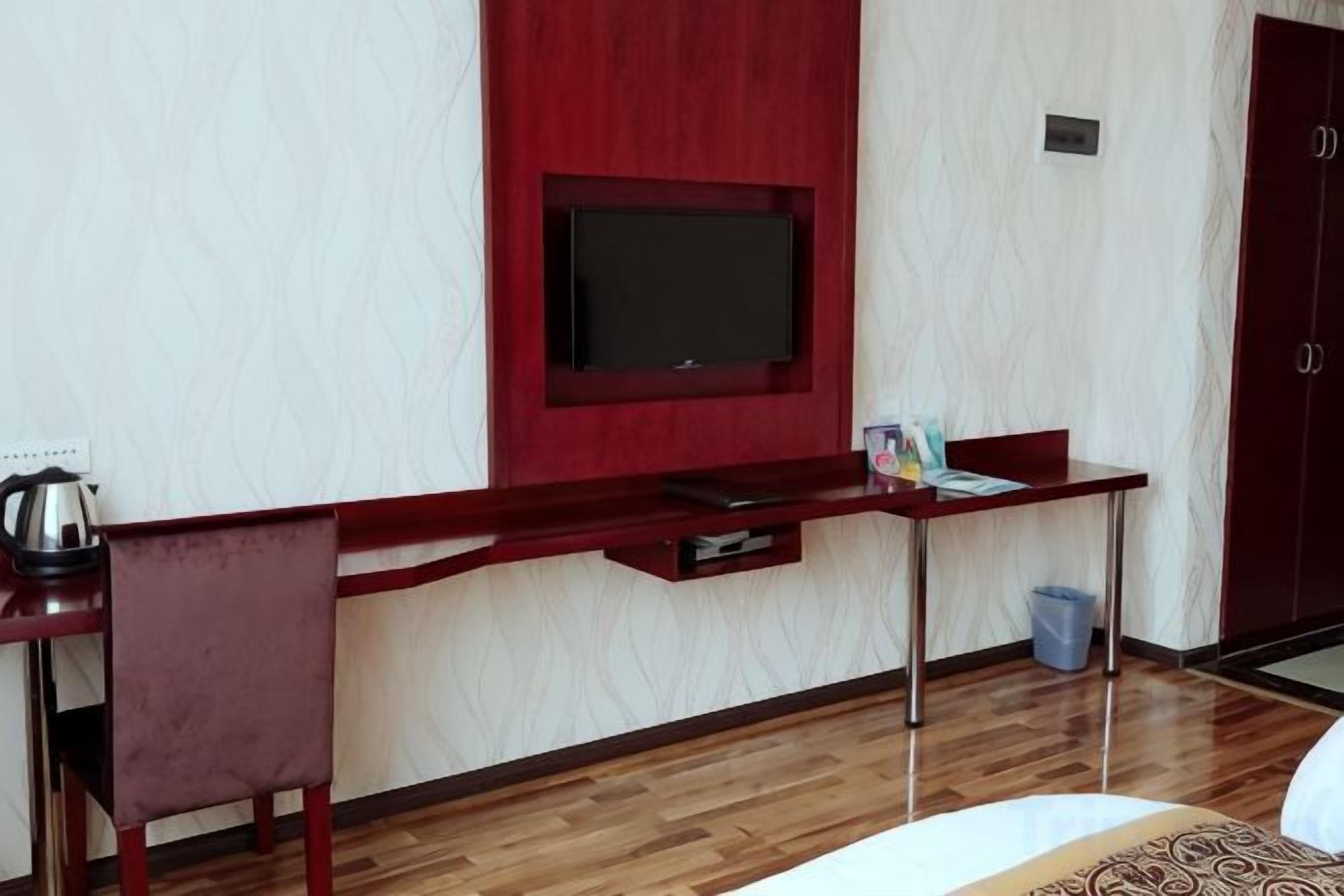 A room with a TV mounted on a red wall, a wooden desk with a chair, a bed, and a door.