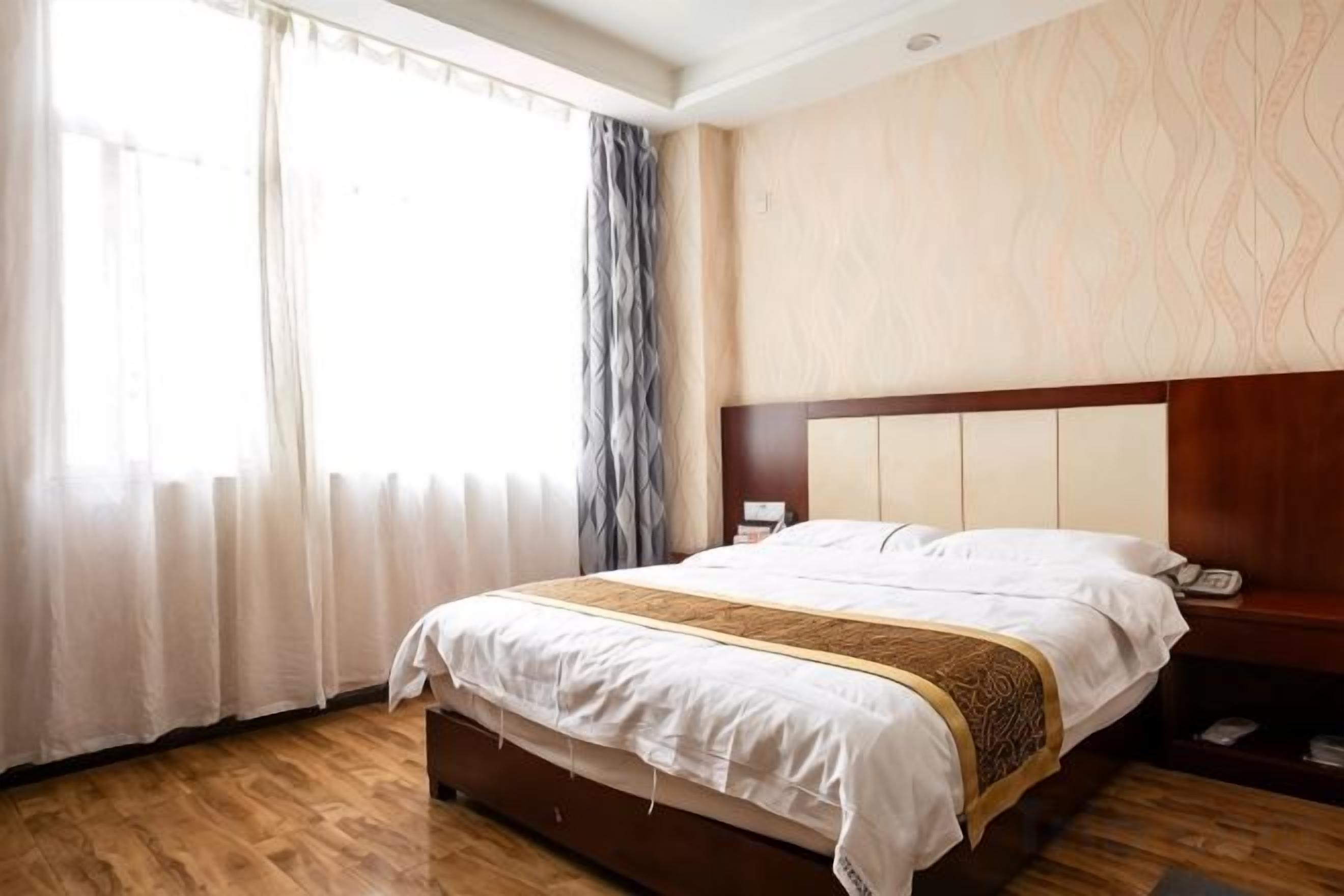 A hotel room with a large bed, a wooden headboard, a bedside table with a lamp, and a window with curtains.