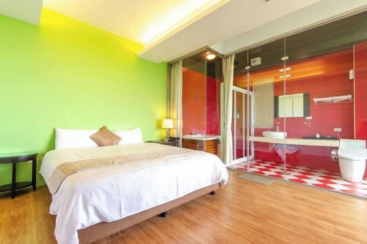 A hotel room with a bed, a nightstand, a lamp, a bathroom with a sink and mirror, and a red and white checkered floor.