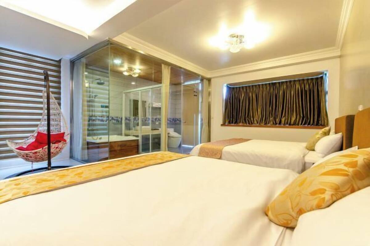 A hotel room with two beds, a glass shower enclosure, a hanging chair, and a view of the bathroom.