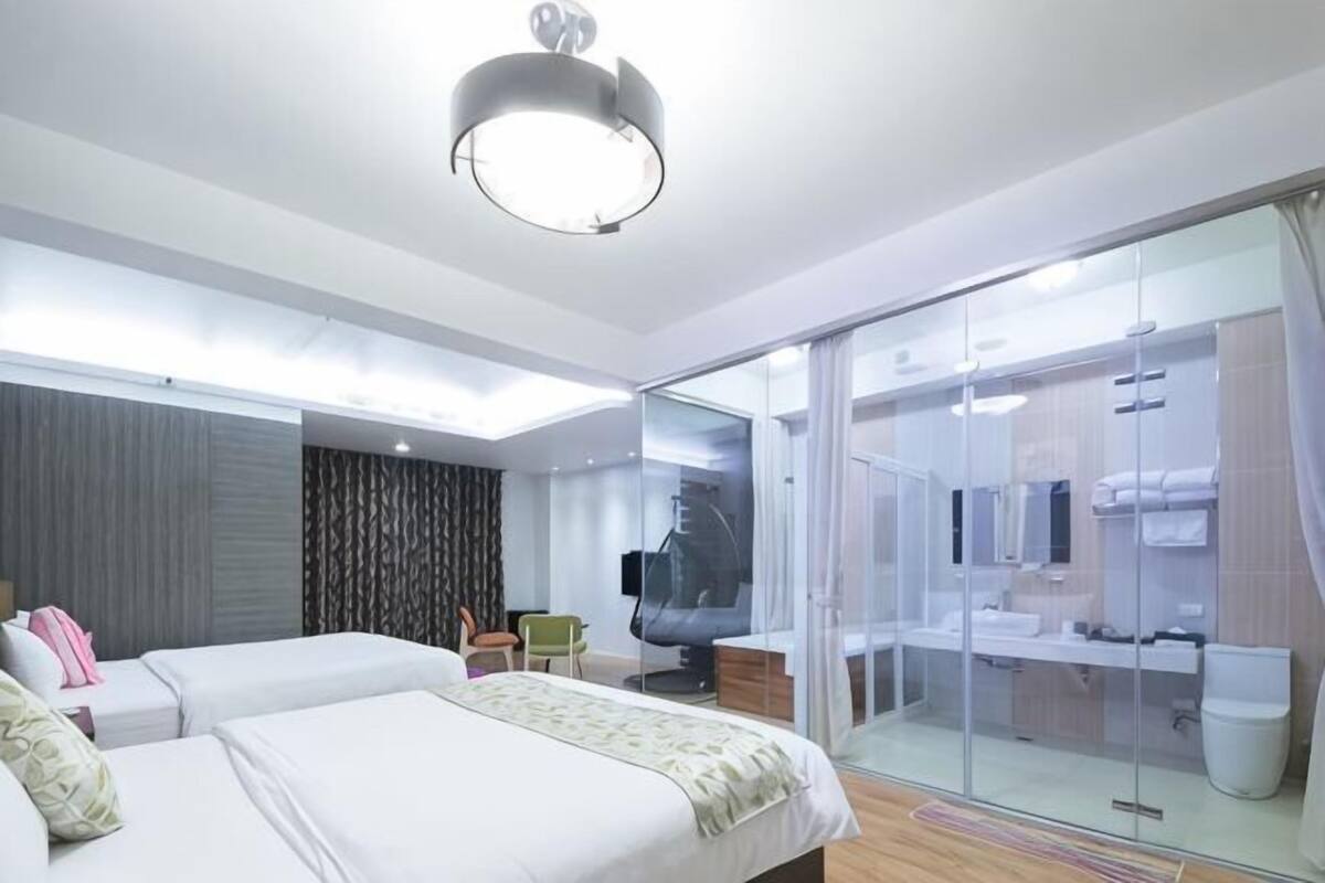 A modern hotel room with two beds, a desk, a chair, a glass shower enclosure, and a bathroom with a sink and toilet.