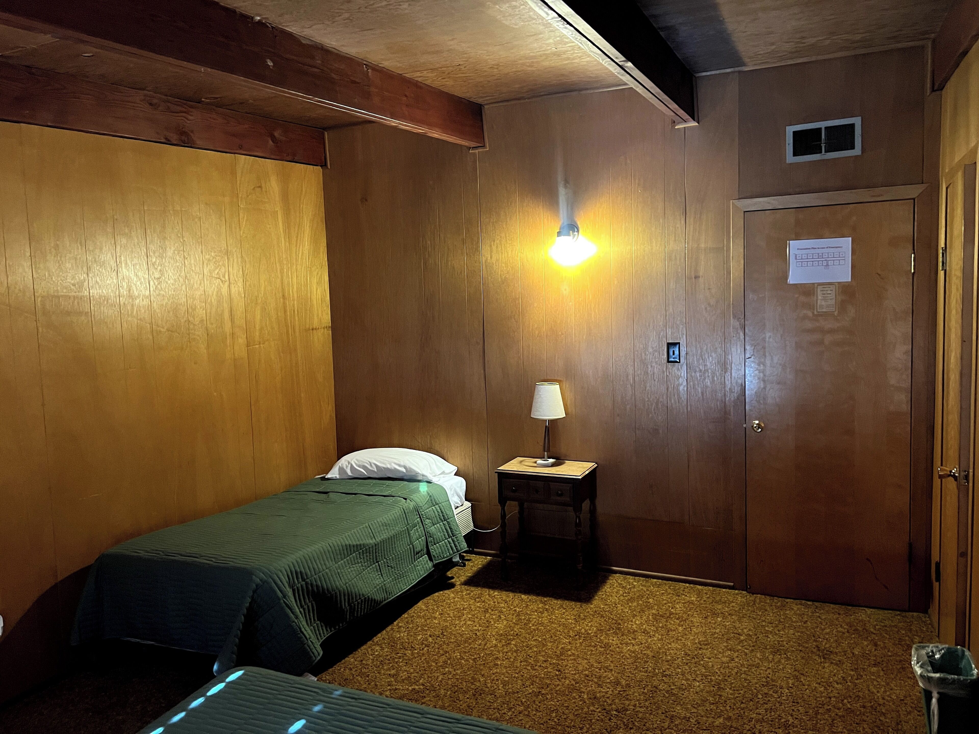 A small, dimly lit room with wooden walls, a single bed, a nightstand with a lamp, and a door with a sign.