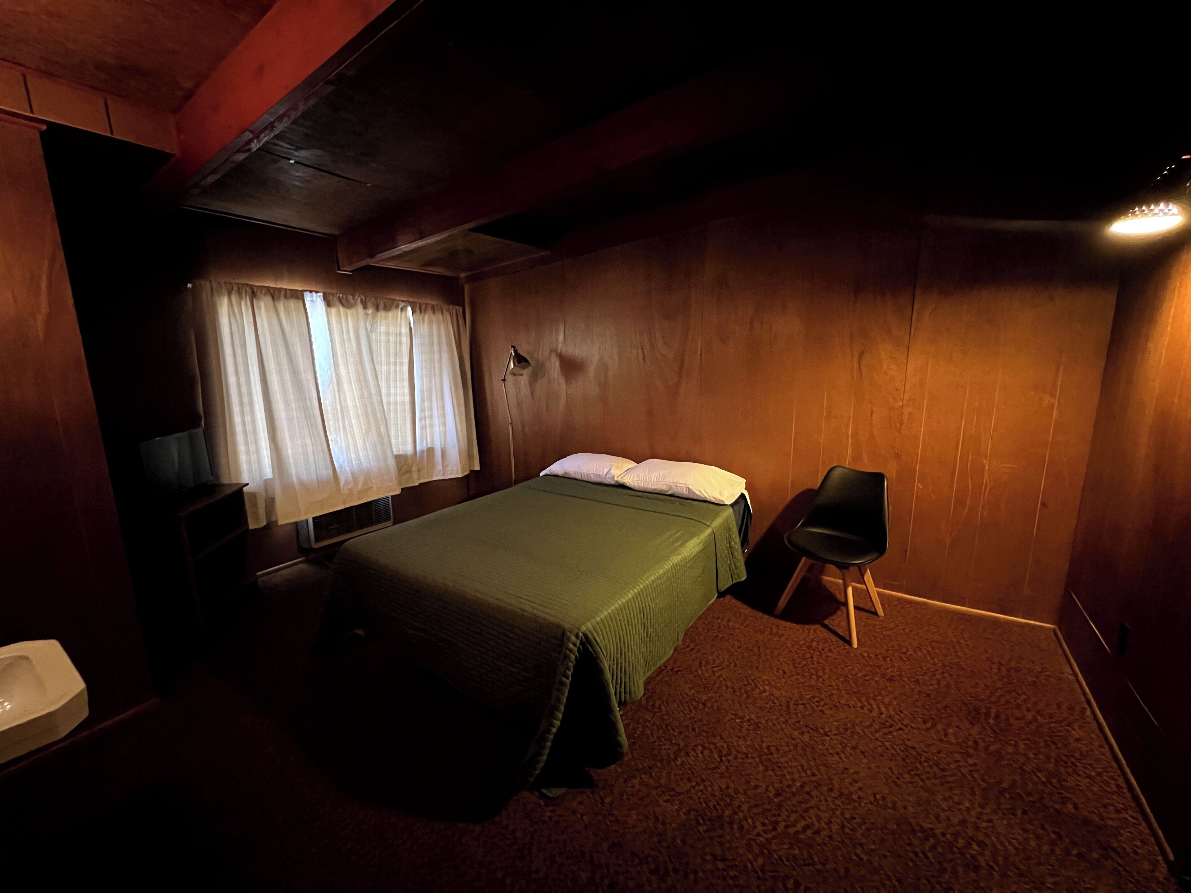 A small, dimly lit room with a single bed, a chair, and a desk with a lamp.