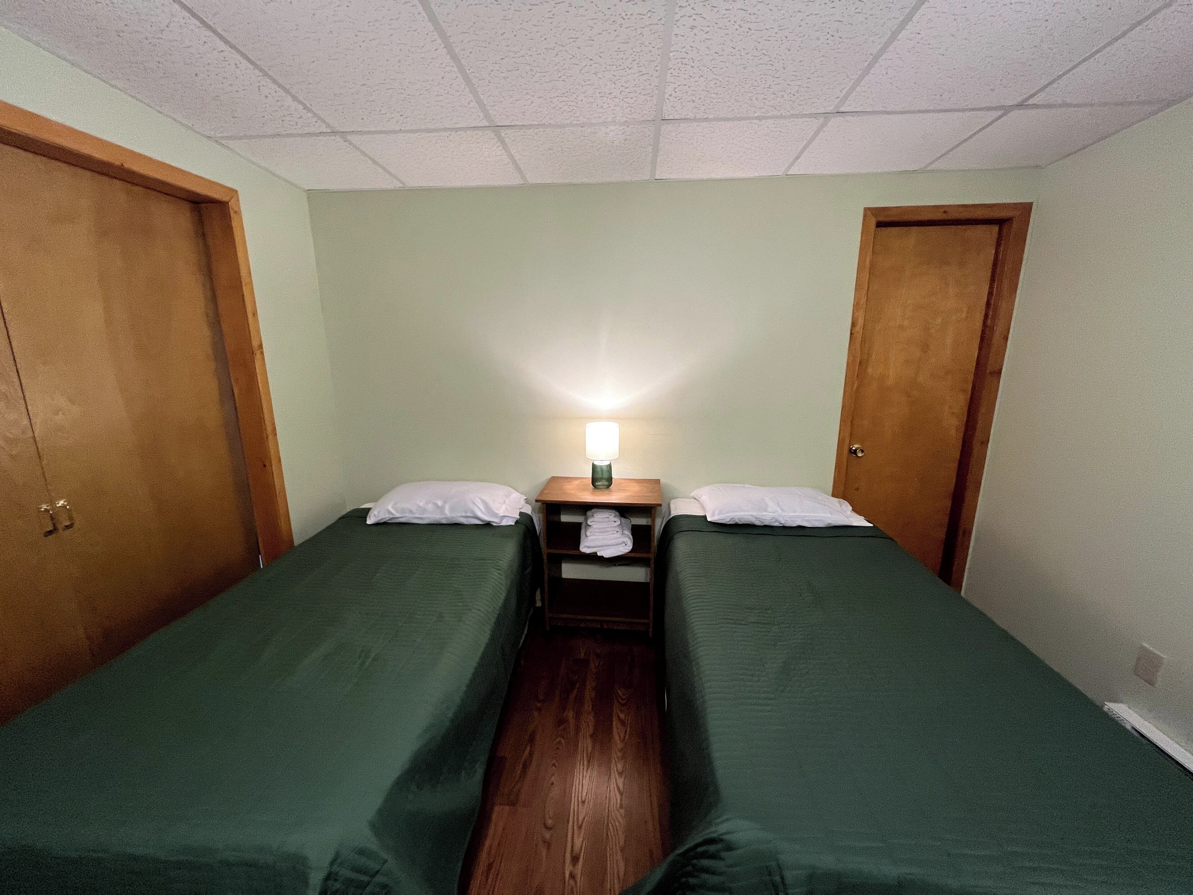 Ein Zimmer mit zwei Einzelbetten, Holztüren, einem Nachttisch mit Lampe und Holzboden.