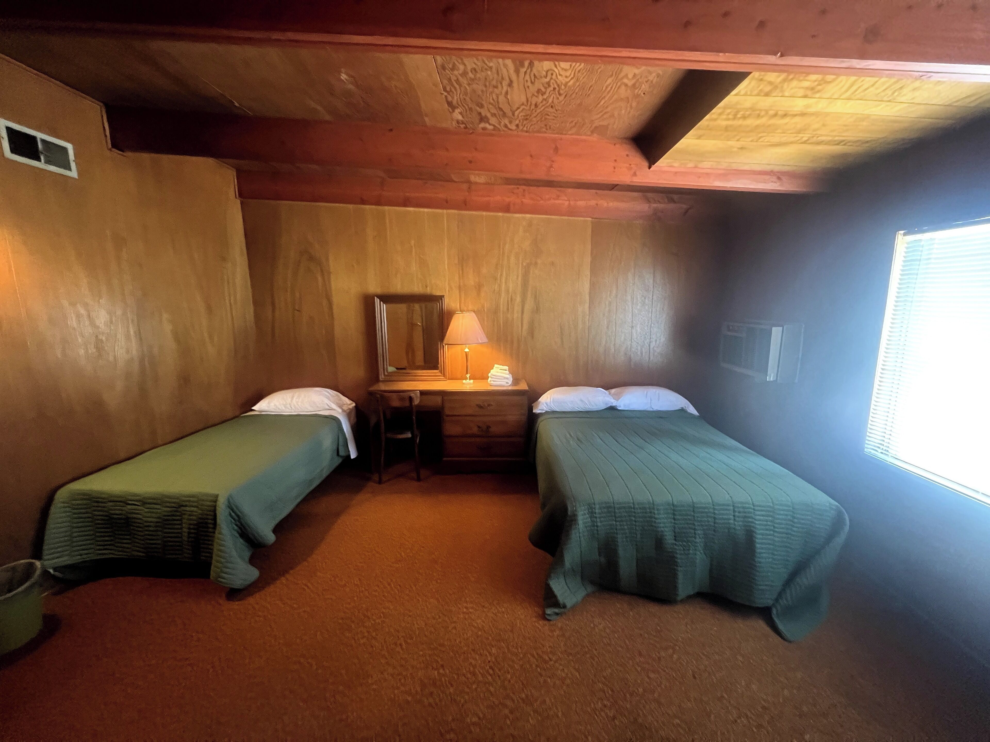 A small, cozy room with two single beds, a wooden desk with a lamp, and a window.