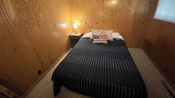 Una habitación con paredes de paneles de madera, una cama con colcha a rayas blanco y negro, y una mesita de noche con lámpara.