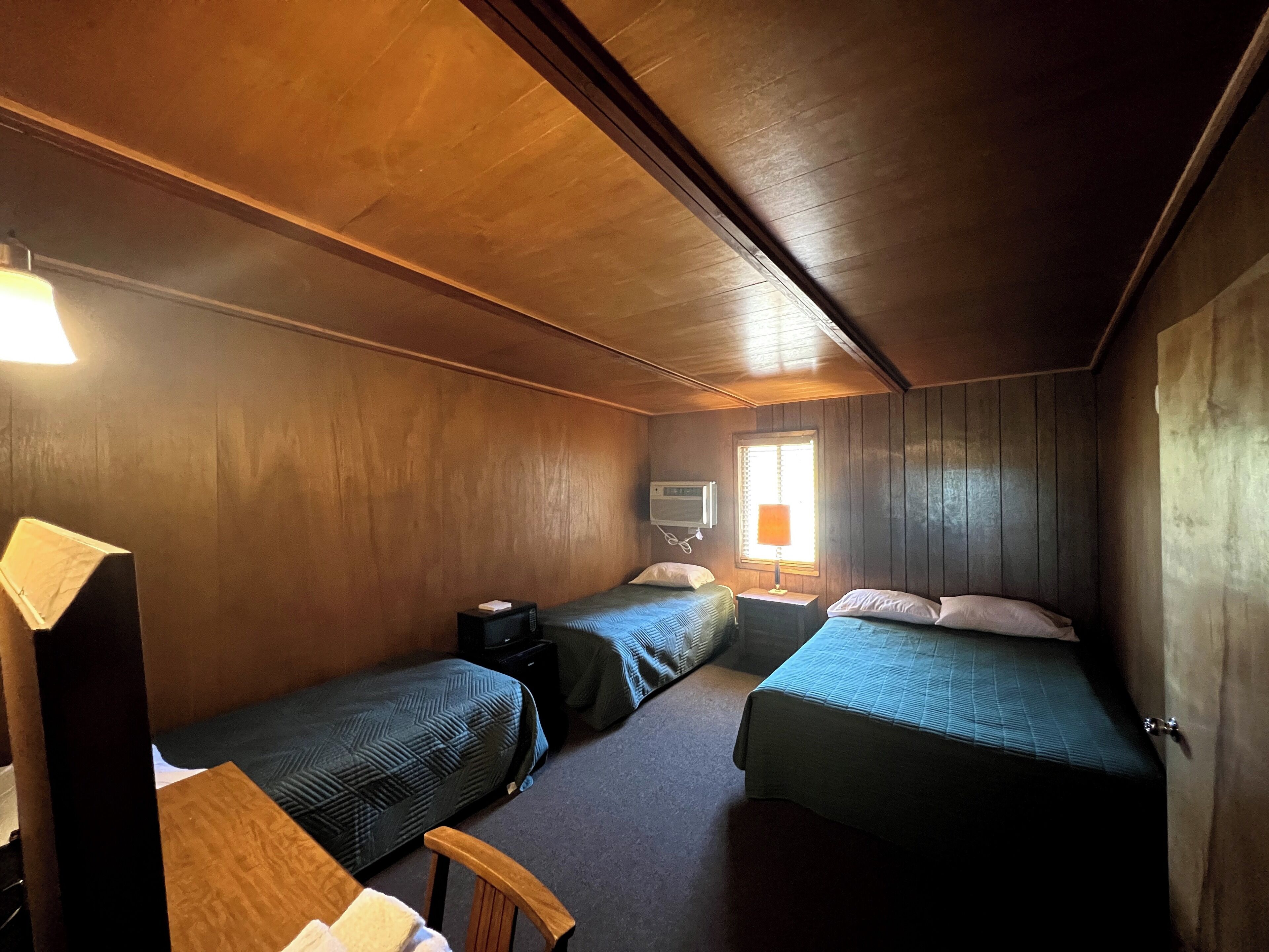A small, cozy room with wooden walls and a sloped ceiling. There are two single beds with matching bedding, a small table with a lamp, and a television on a stand.